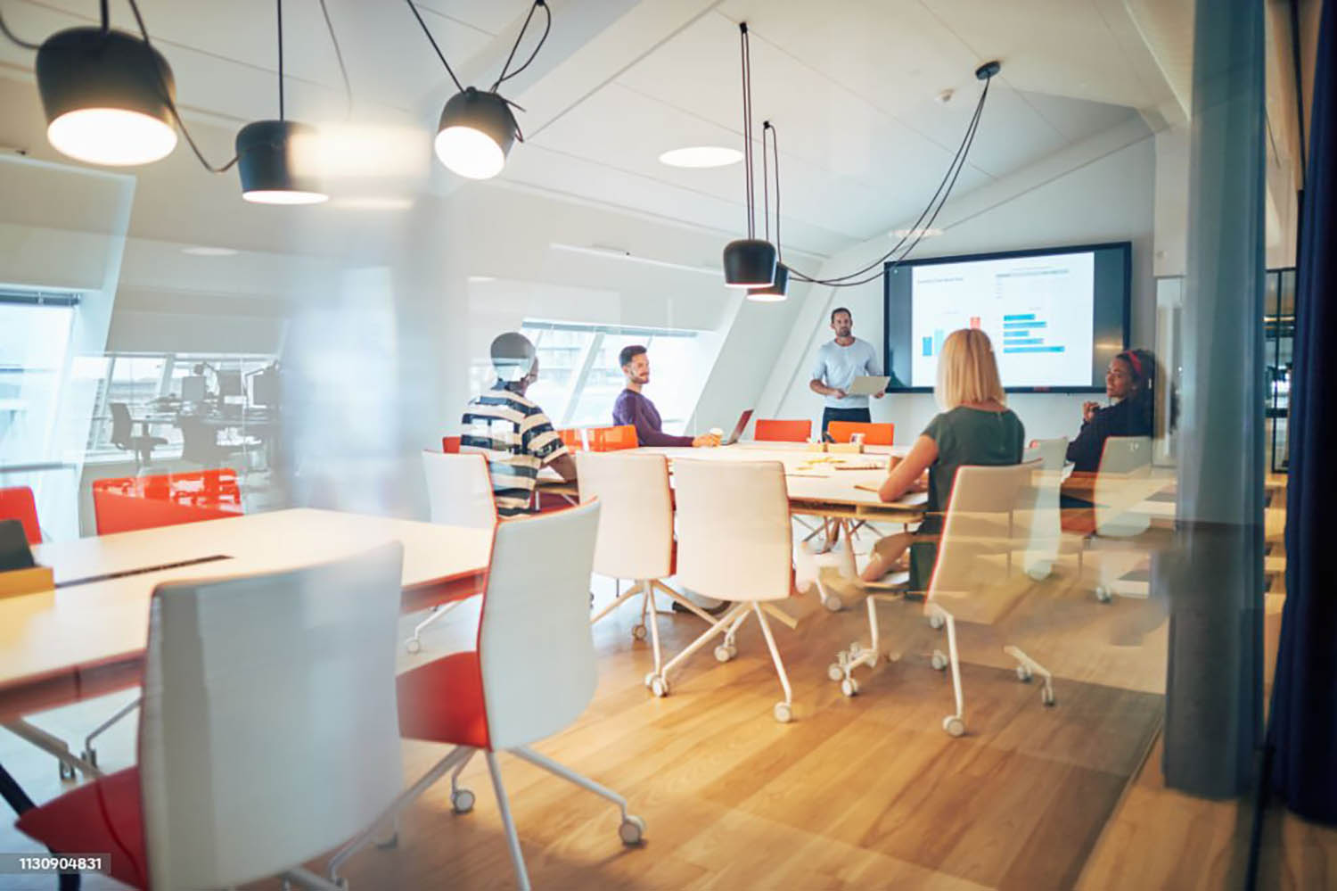 Manager discussing graphs with a diverse group of colleagues during a monitor presentation inside of a glass office